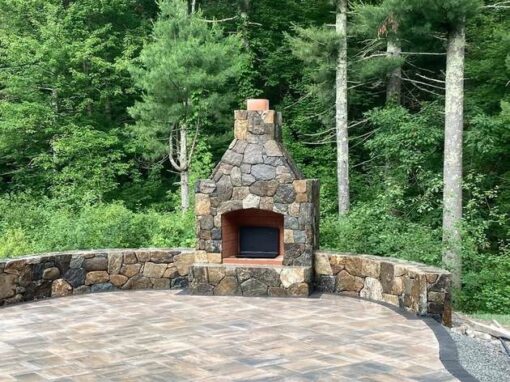 Stone fireplace and curved seat walls at the edge of a wooded paver patio