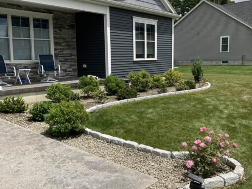 Foundation shrubs and roses in a stone-edged bed along a gray ranch