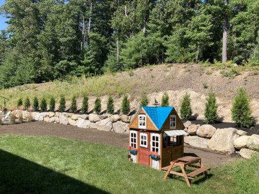 Boulder wall with arborvitae behind a play area