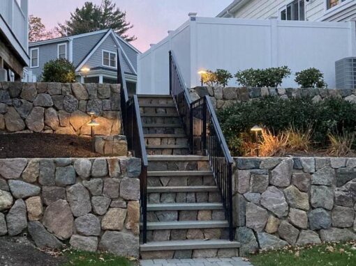 Lit stone steps with a black railing through a two-tier fieldstone wall